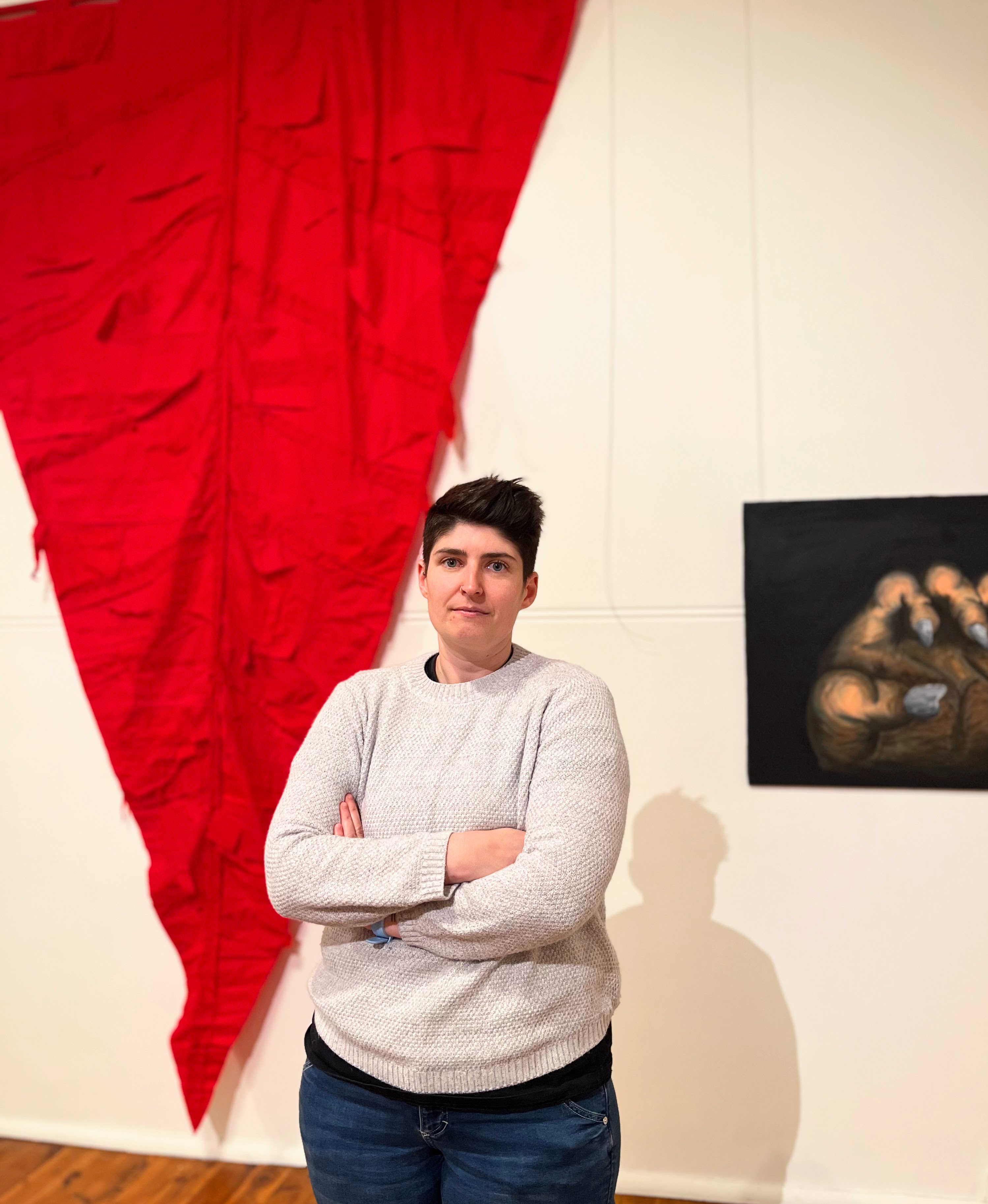 A woman with her arms crossed stands beside a large red piece of fabric pinned to a wall. 