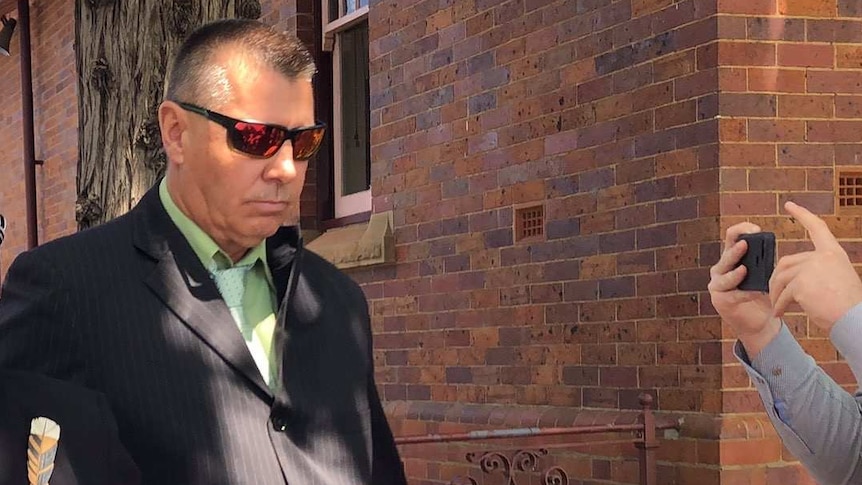 man with sunglases, brown suit and green shirt walking outside a building