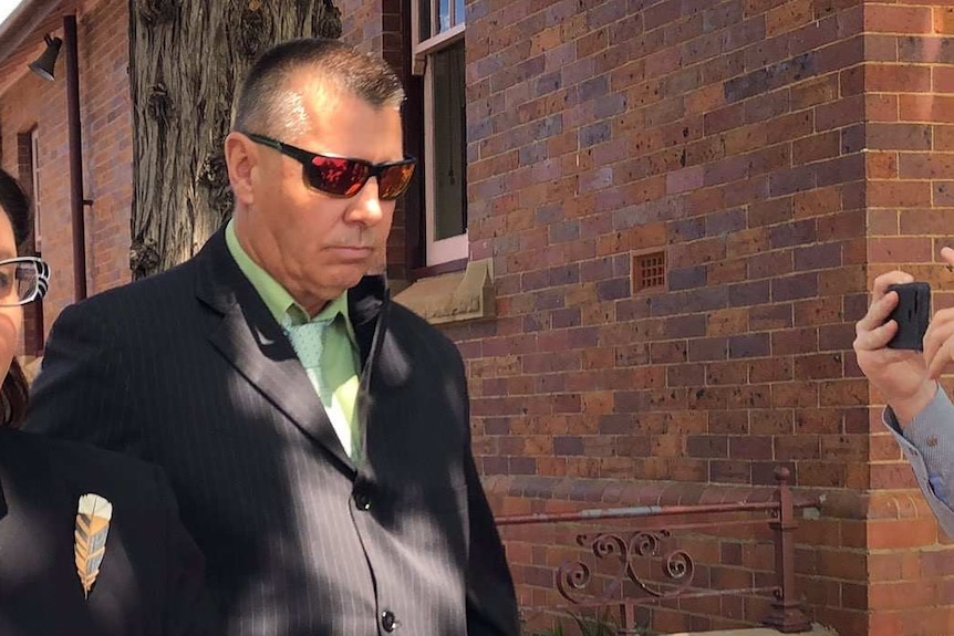 man with sunglases, brown suit and green shirt walking outside a building