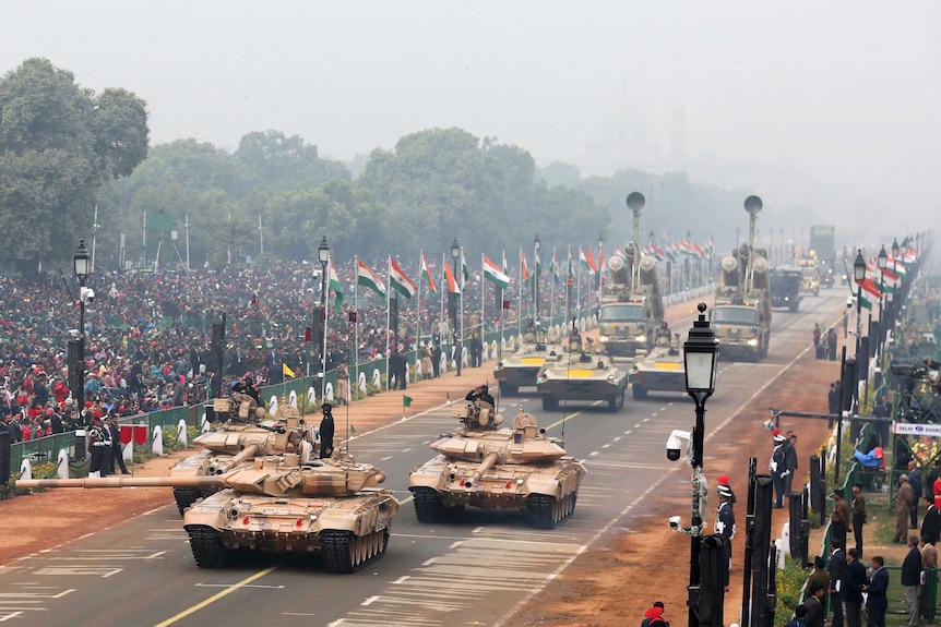A wide-view shows Indian army tanks and other military equipment rolling along a street with crowds watching on.