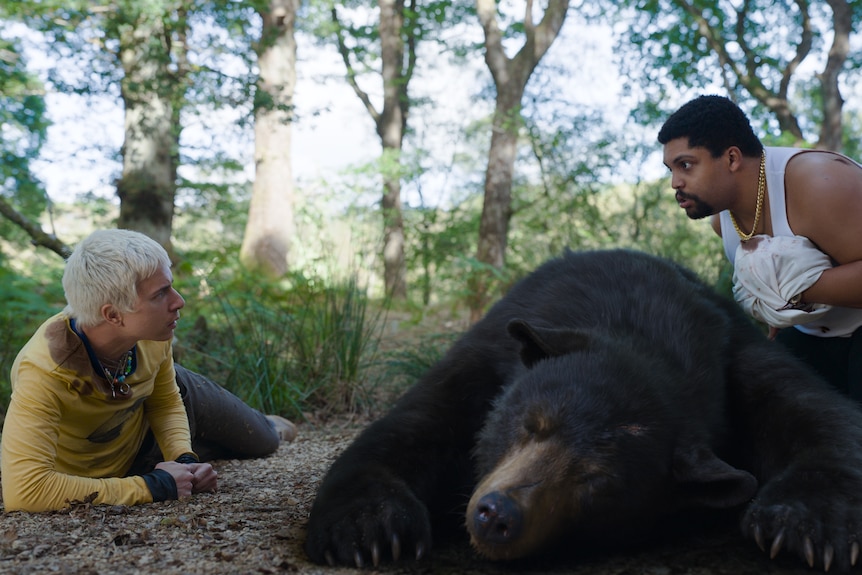 A white man with short white blonde hair is lying next to an unconscious bear. A black man with short dark hair kneels beside.