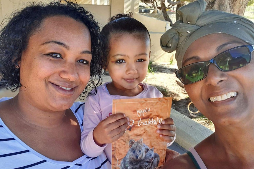 A young girl holds a book as she is held between two smiling women in headshot.