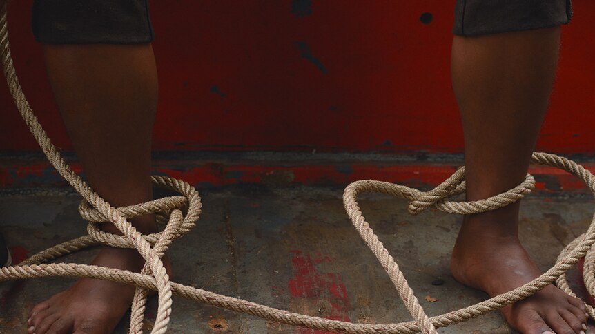 Close-up of barefooted person with sand coloured natural fiber rope loosely tied around ankles on paint faded boat surface.