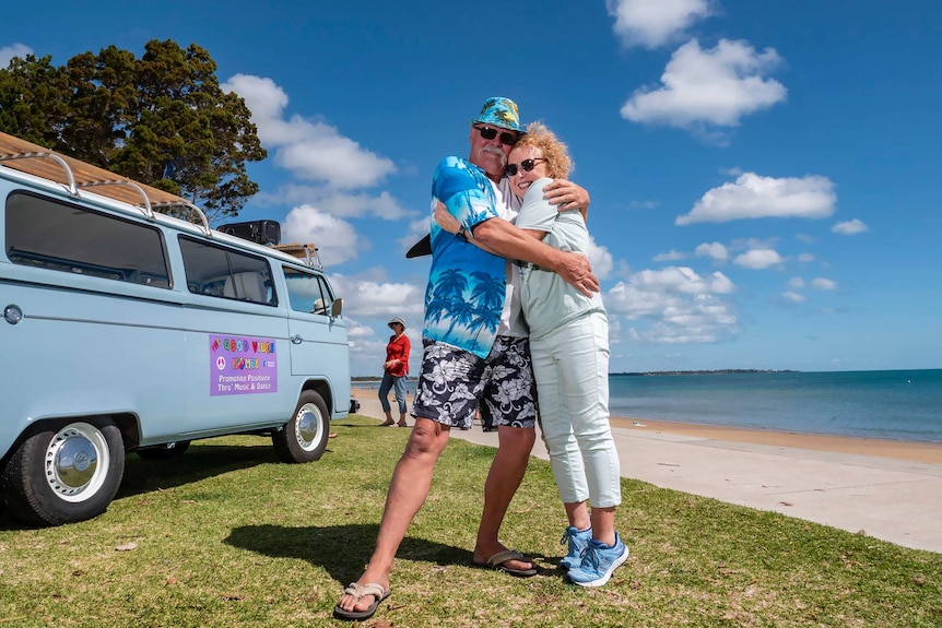 A man and woman hug in front of a van on the seaside.