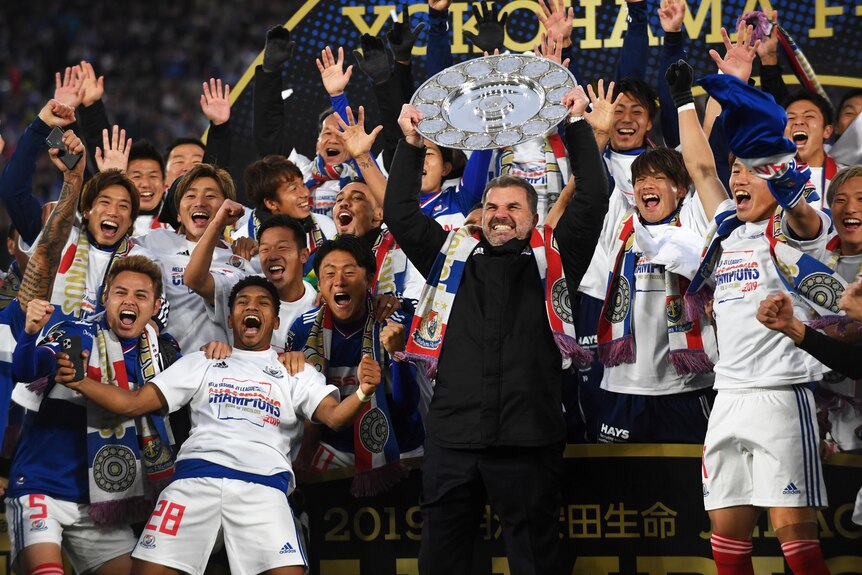 Ange Postecoglou lifts a round sliver plate as men in white shirts smile and scream in joy