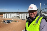A man in a high vis vest and helmet overlooks a worksite with large storage vats for wine.