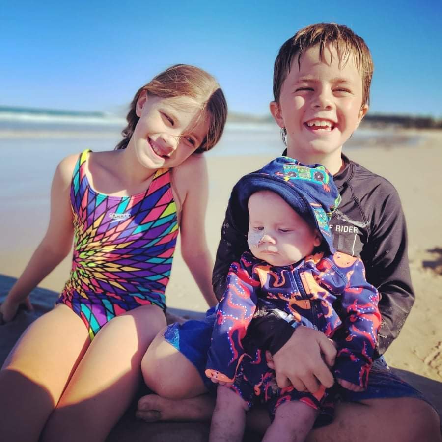 Baby Austin and his siblings at the beach wearing bathers.