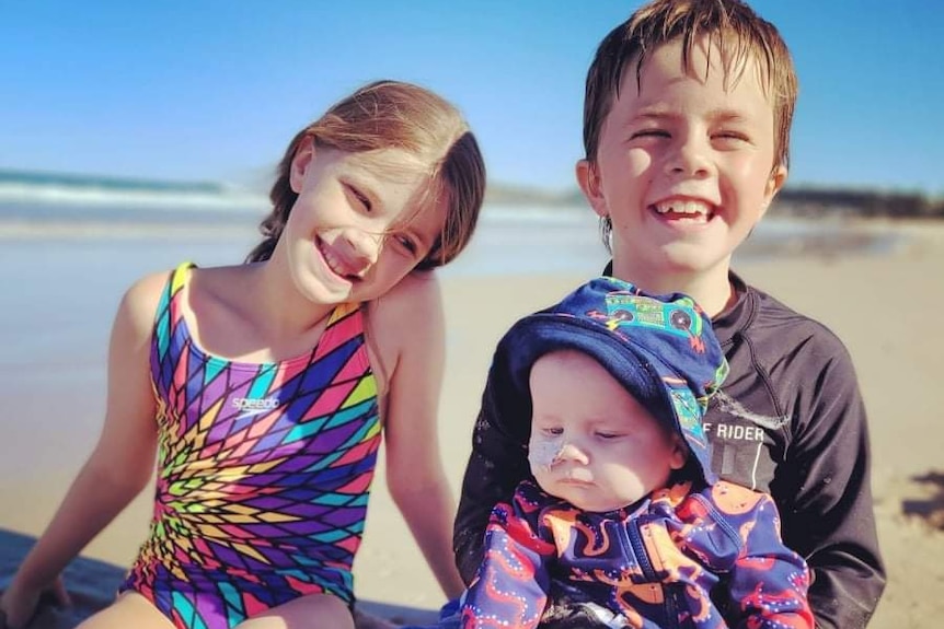 Baby Austin and his siblings at the beach wearing bathers.