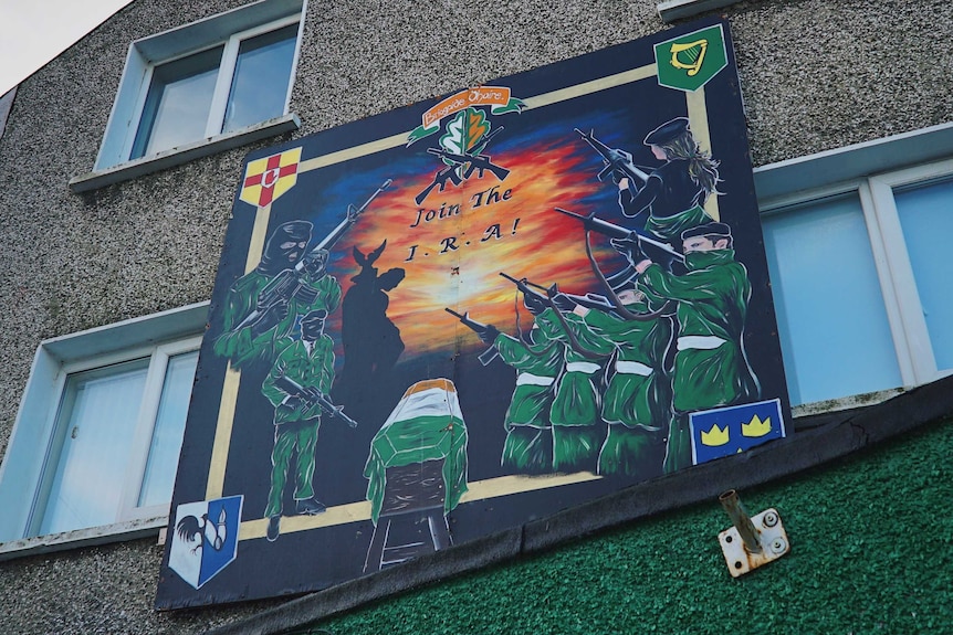 A mural shows IRA soldiers in green holding guns with a coffin shrouded in Irish flag and "join the IRA" over bright sunset.