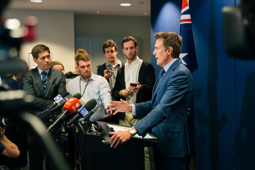 A group of mainly male reporters look at Mr Porter as he speaks at a podium.