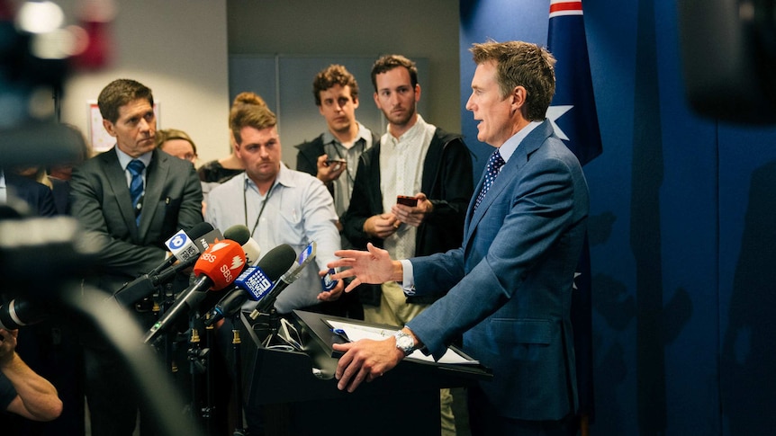 A group of mainly male reporters look at Mr Porter as he speaks at a podium.