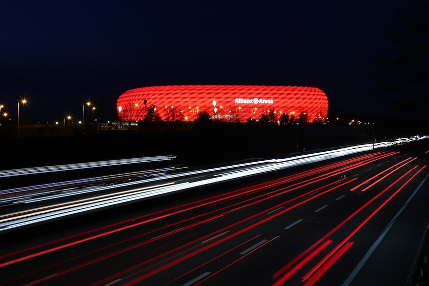 At night time, cars drive past a Munich football stadium lit up in red.