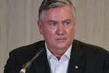 Eddie McGuire sits wearing a black suit and white, open necked shirt, with a microphone in front of him