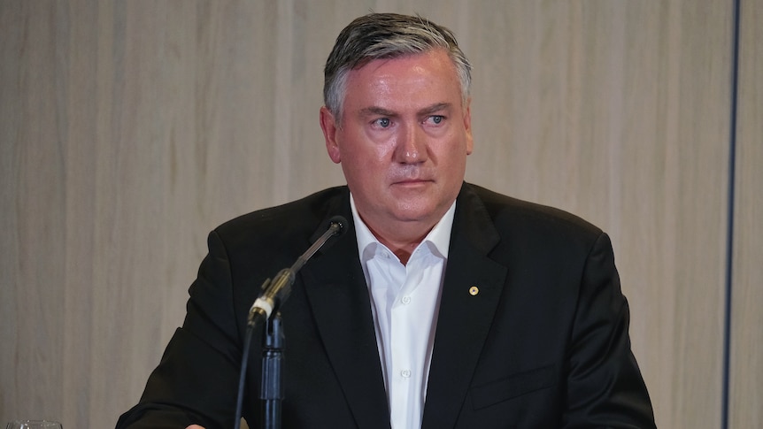 Eddie McGuire sits wearing a black suit and white, open necked shirt, with a microphone in front of him