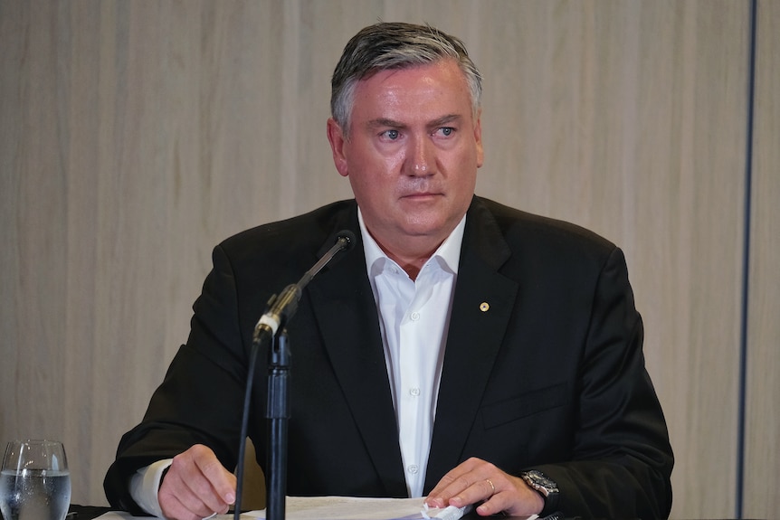 Eddie McGuire sits wearing a black suit and white, open necked shirt, with a microphone in front of him