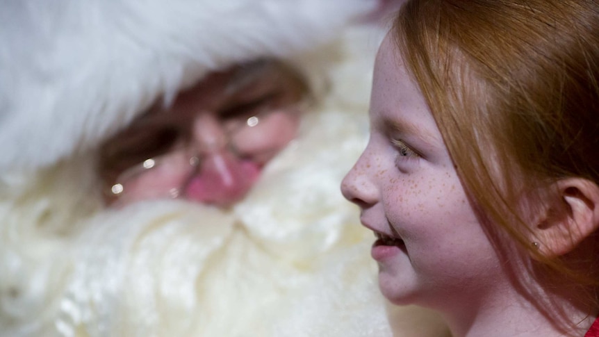 A little girl smiles as Santa's big face and beard is out of focus behind her.