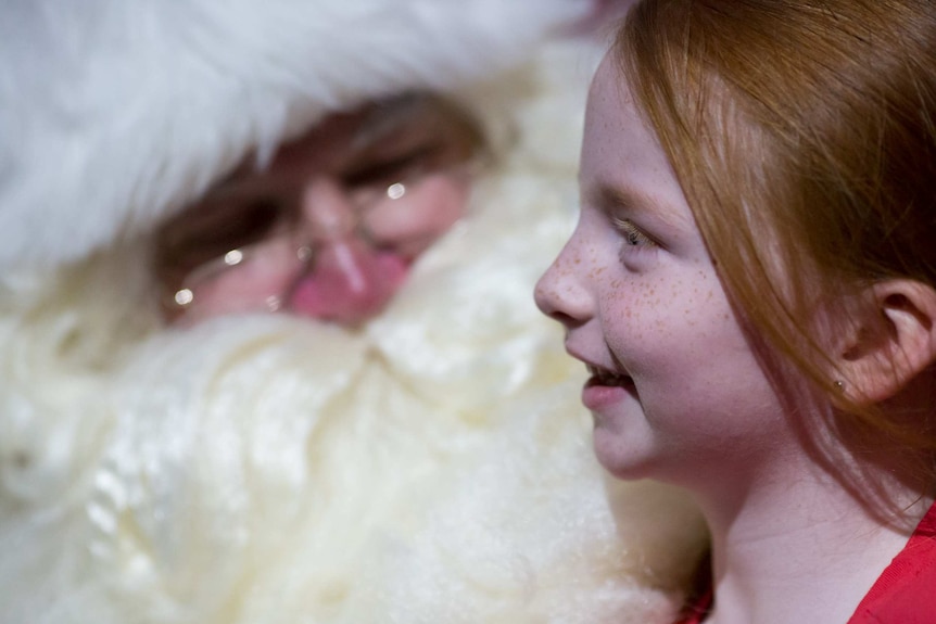 A little girl smiles as Santa's big face and beard is out of focus behind her.