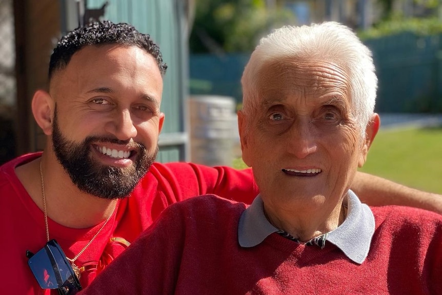 A young maori man with short curly dark hair sits beside his elderly grandfather with grey hair, both smiling and wearing red