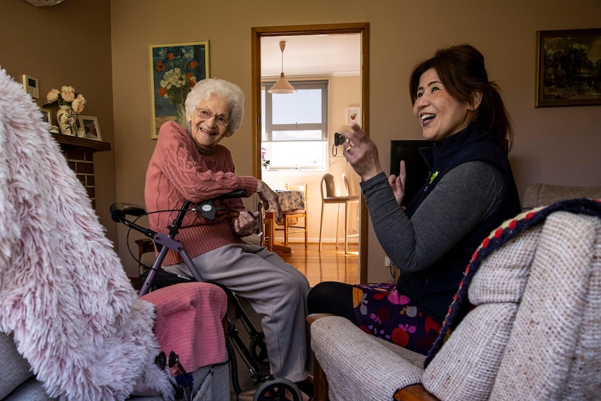 An elderly woman and another woman chat and laugh in a living room.