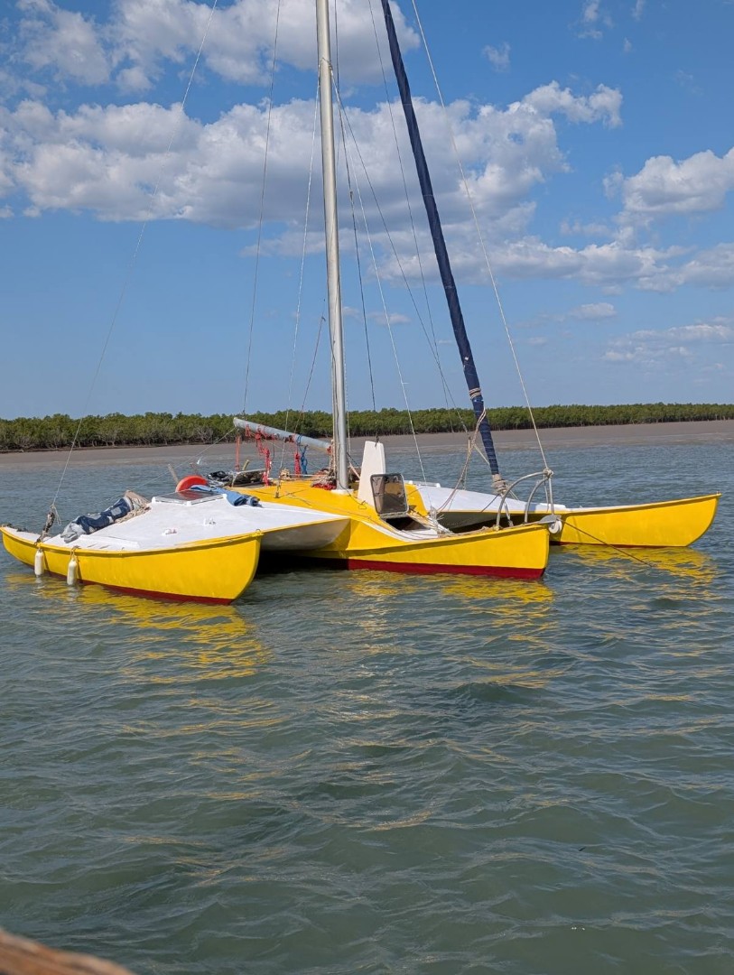 A trimaran boat with yellow paint on its sides and red on its underbelly floating in the water just off the coast.