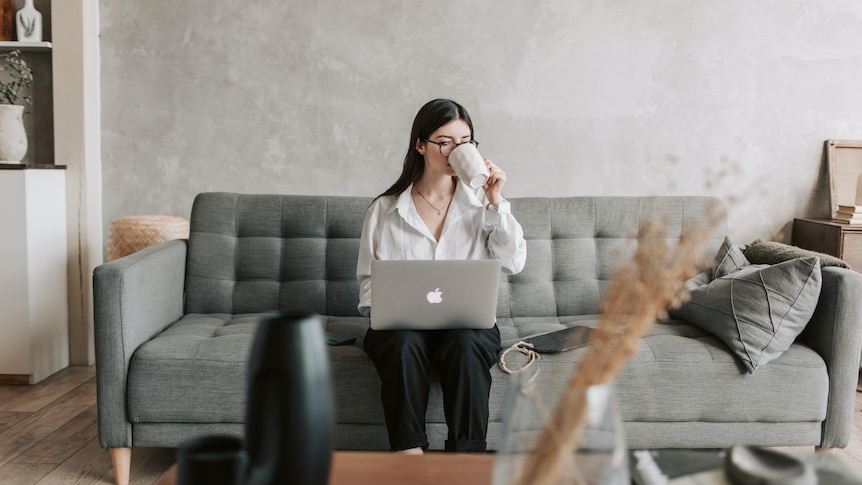 Woman with cup in hand sits on couch with laptop on lap at home