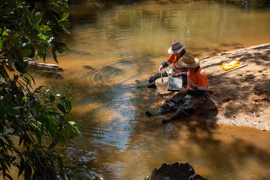 Two people with akubra hats on sit on a sandy bank on the edge of a murky creek 