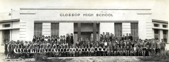 A black and white photo of a school building with rows of students sitting out the front.