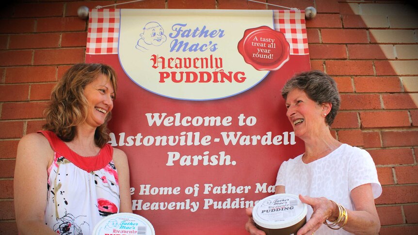 Two women hold packaged pudding in front of a sign that reads 'Father Mac's heavenly pudding'.