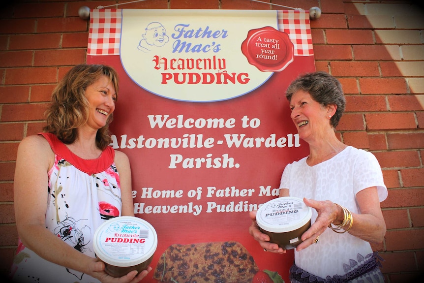 Two women hold packaged pudding in front of a sign that reads 'Father Mac's heavenly pudding'.