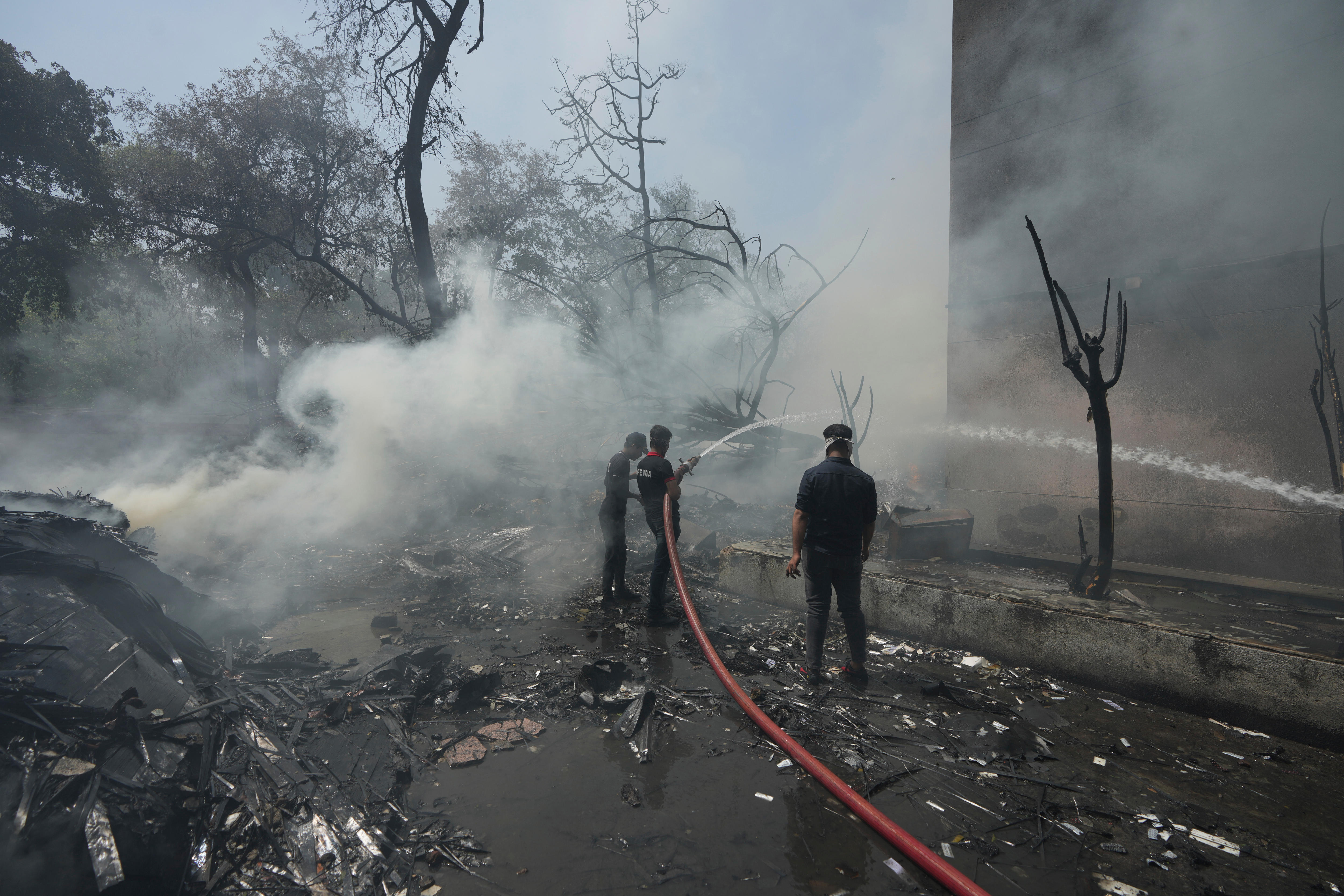 Men standing in a charred street surrounded by blackened tree stumps and white smoke plumes while using a red fire hose