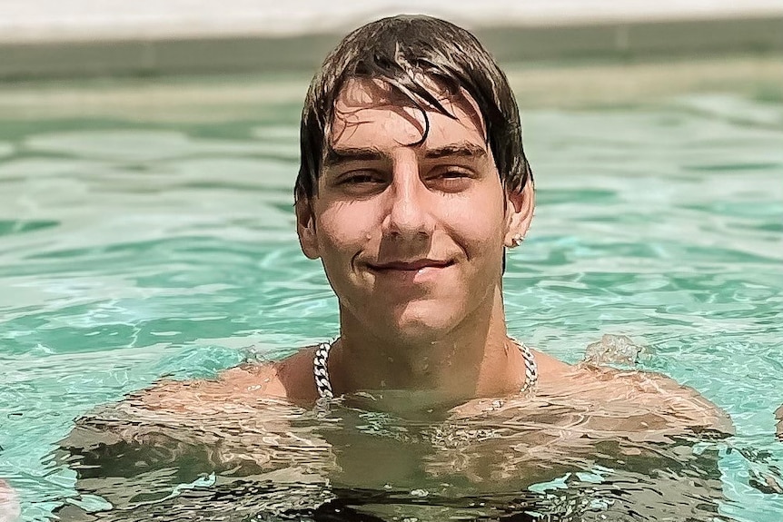 a young boy smiling at the camera while in a pool wearing a silver chain around his neck
