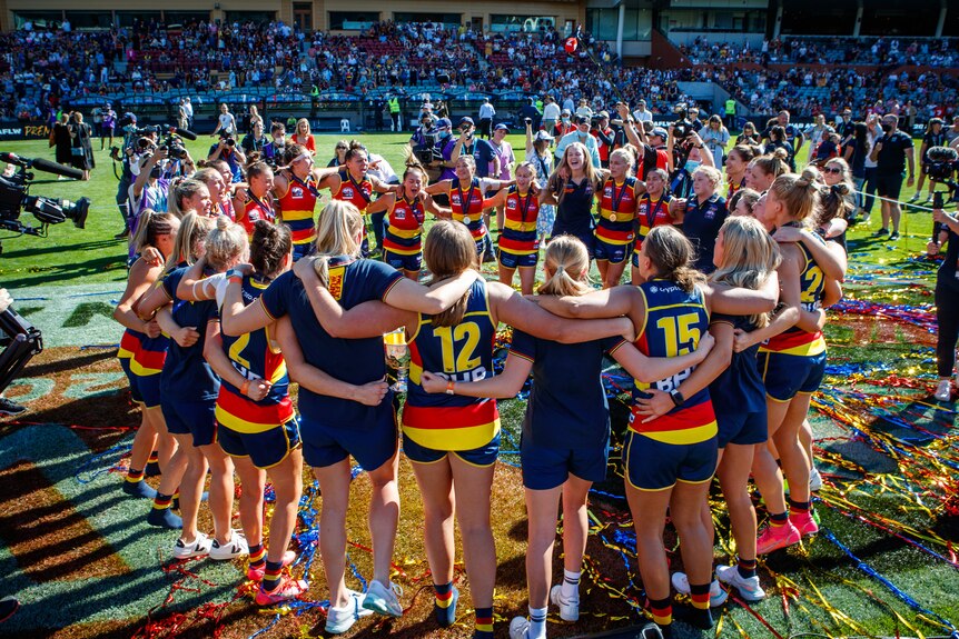 The Adelaide Crows AFLW team stand in a circle, arm in arm.