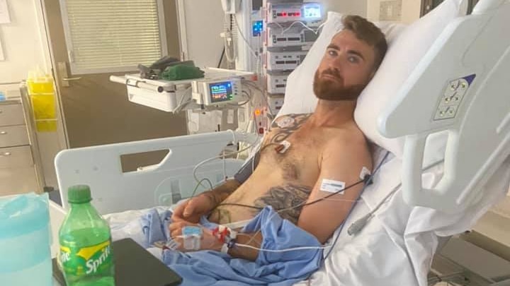 A young man, bearded and shirtless, hooked up to monitoring equipment in in a hospital bed.