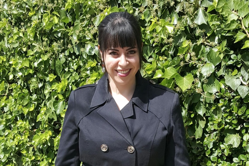 Dassi Erlich wears black and smiles while standing in front of a shrub.