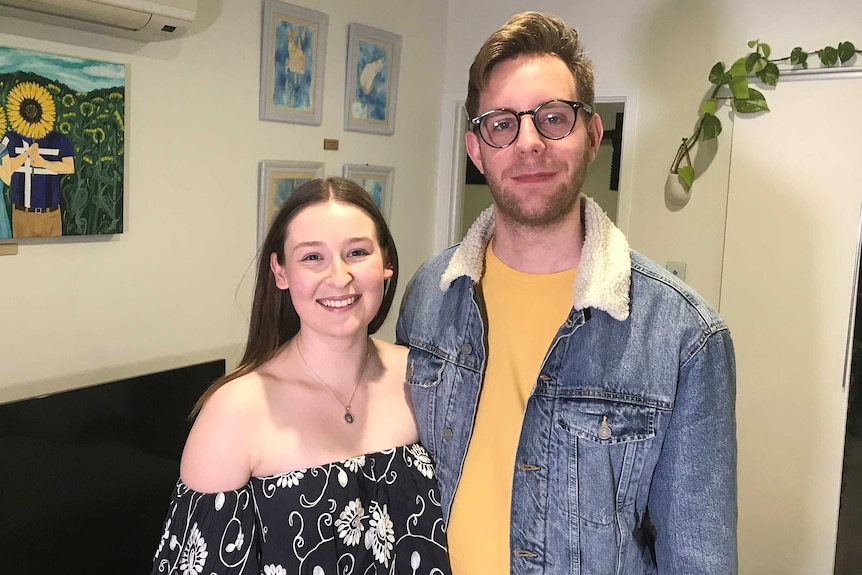 Iliria and Tom Stenning standing inside their apartment in front of a wall full of paintings.