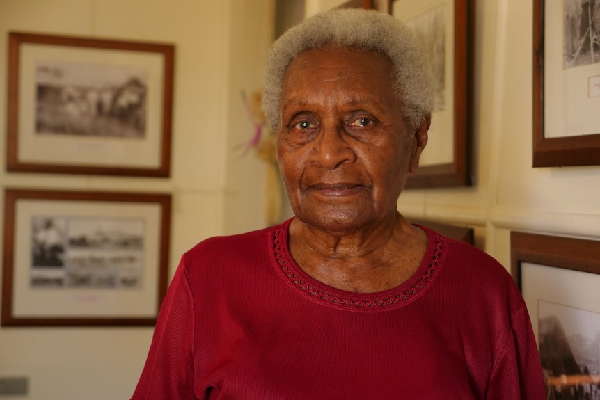 Aunty Doris straight-faced, red shirt, historic images on wall behind.