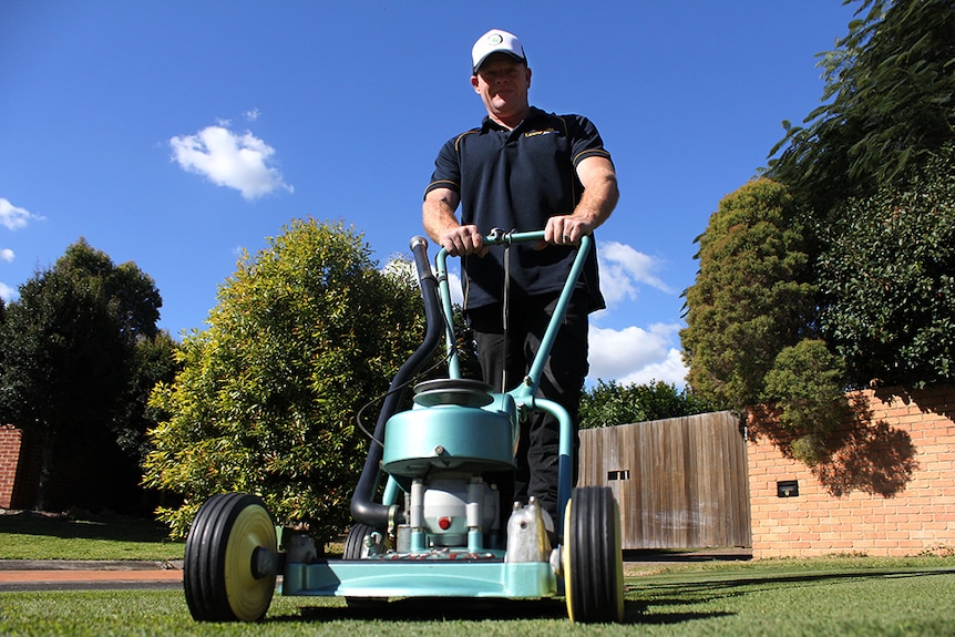 A man in jeans and a polo shirt pushing an old style, two-stroke, Victa lawn mower.