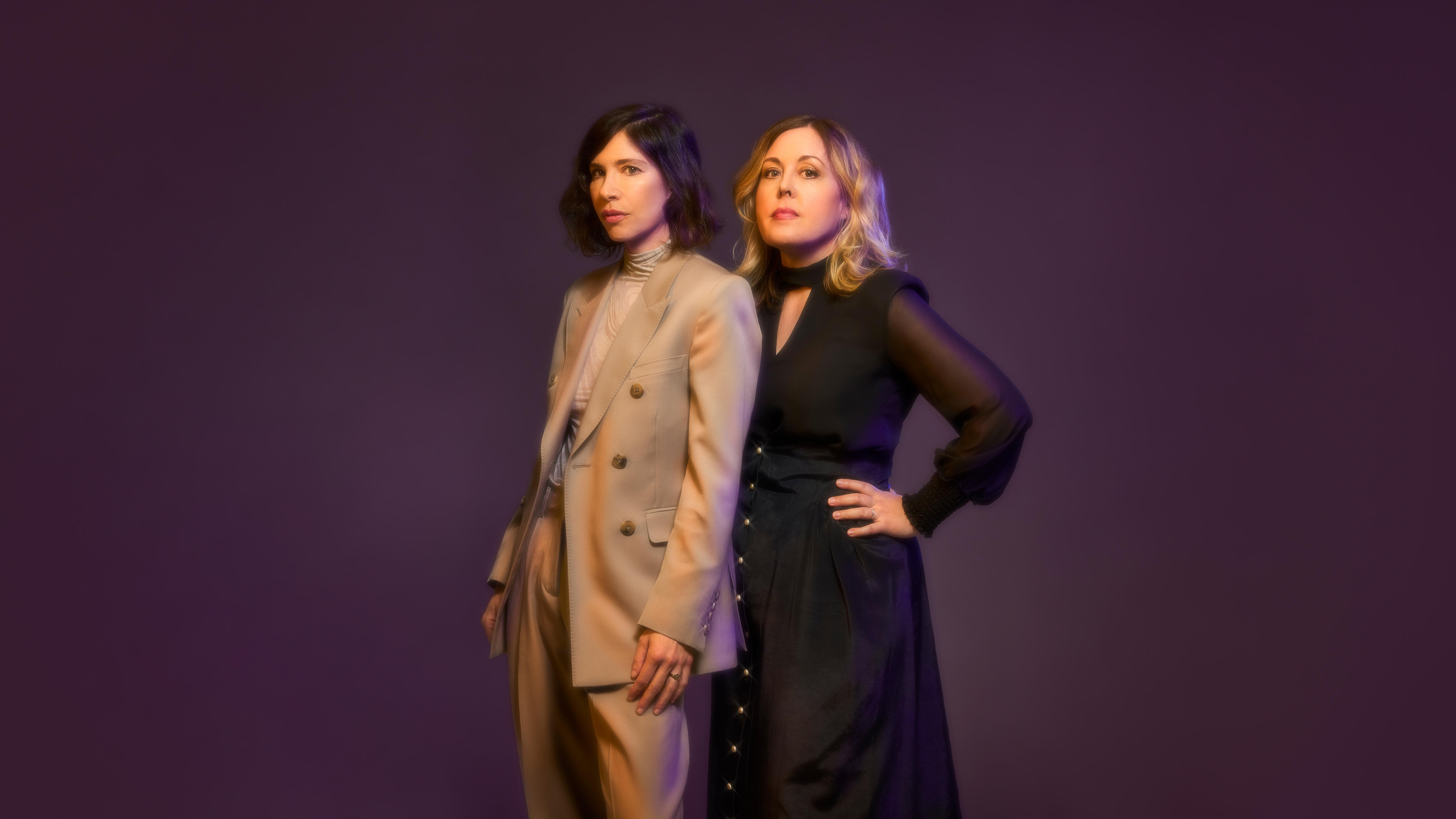 Carrie Brownstein and Corin Tucker stand side-by-side against a purple background