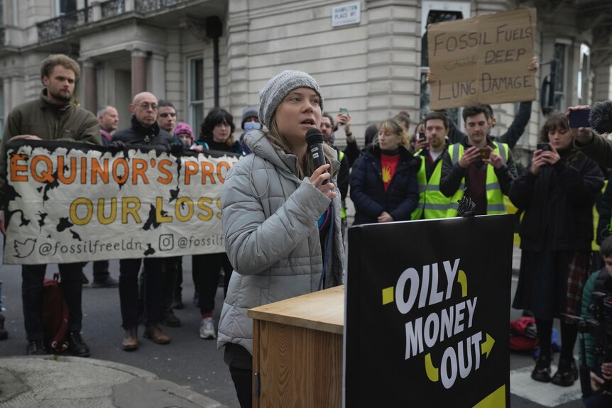 Greta Thunberg speaking into a microphone with protesters surrounding her and a sign reading "Oily Money Out" in front of her