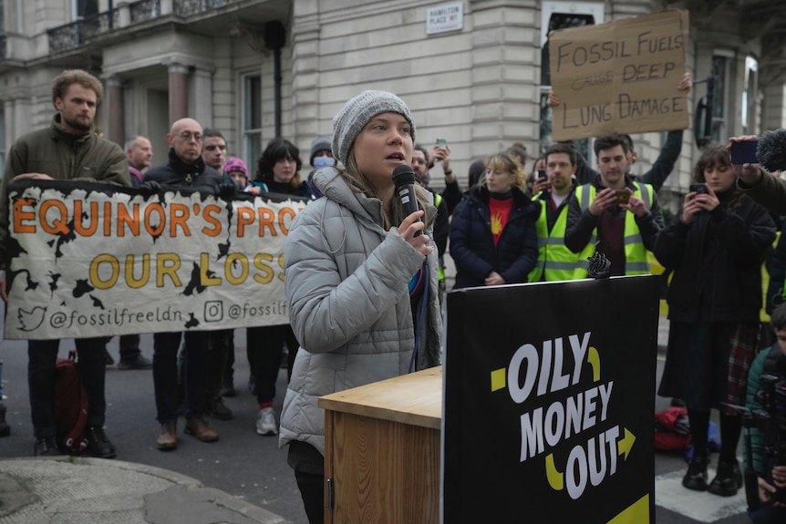 Greta Thunberg vorbește într-un microfon în timp ce protestatarii o înconjoară și pe un banner scrie "Banii uleiosi au iesit" In fata ei