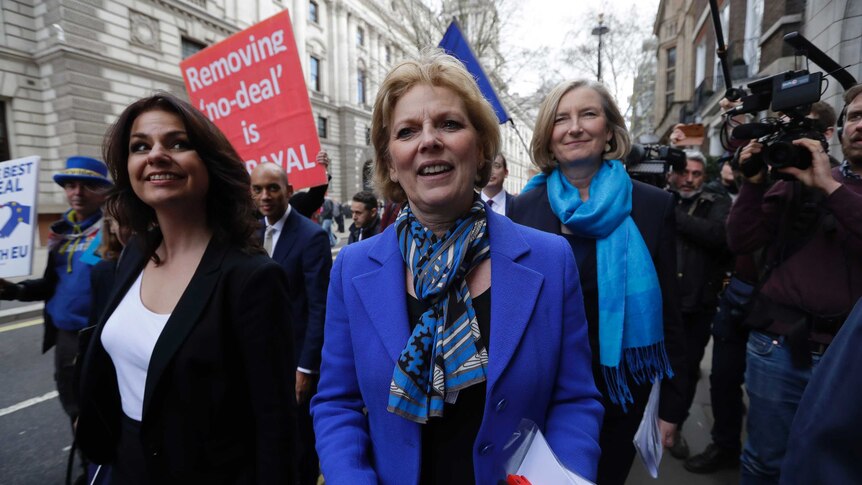 Three women dressed in blue walk in the street as supporters follow behind.