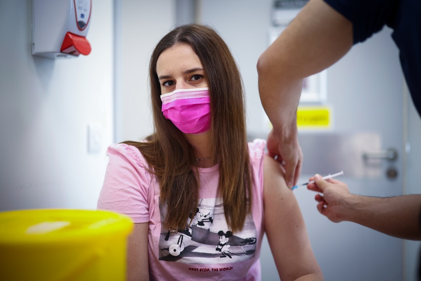 A woman in a pink face mask gets a needle injected into her shoulder 