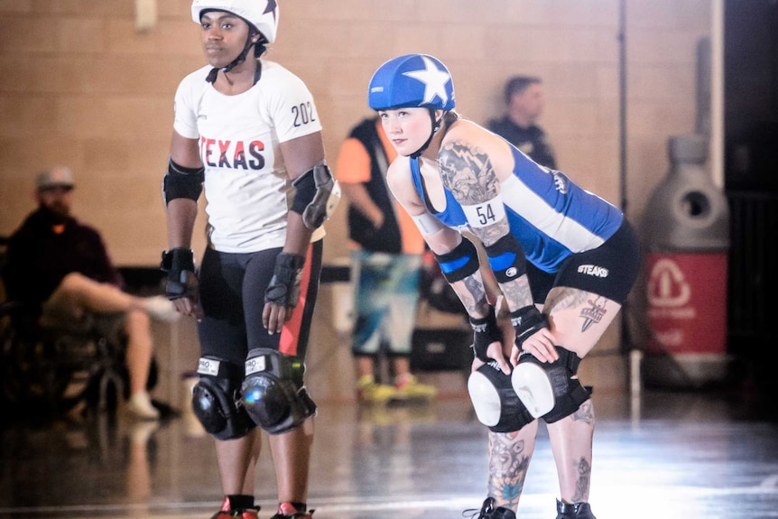 Two women in roller skates, knee and elbow pads and helmets, one in white, one in blue.