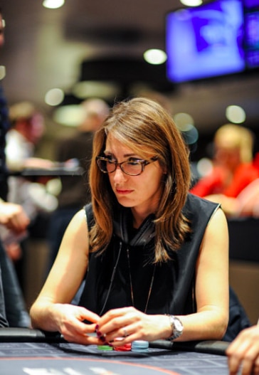 Alex O'Brien, with glasses, shoulder-length brown hair and dark blouse, sits at poker table holding chips, looking focused. 