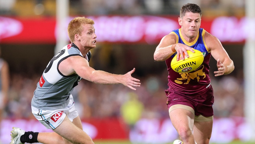 A Brisbane Lions AFL player looks down at the ball ready to drop it on his boot, while a defender scrambles to stop him.