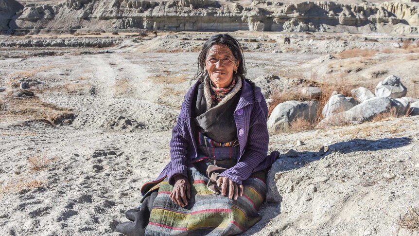 An elderly woman sits outside in the mountains in traditional dress