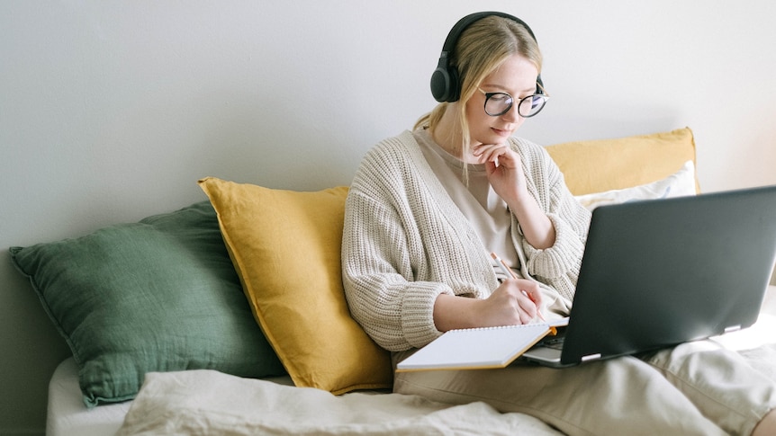 A woman with blonde hair and glasses sits on some cushions in front of a laptop, with headphones on