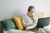 A woman with blonde hair and glasses sits on some cushions in front of a laptop, with headphones on