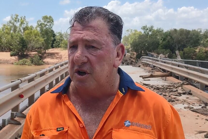 A man in a high-vis orange shirt speaks in front of a damaged bridge. 