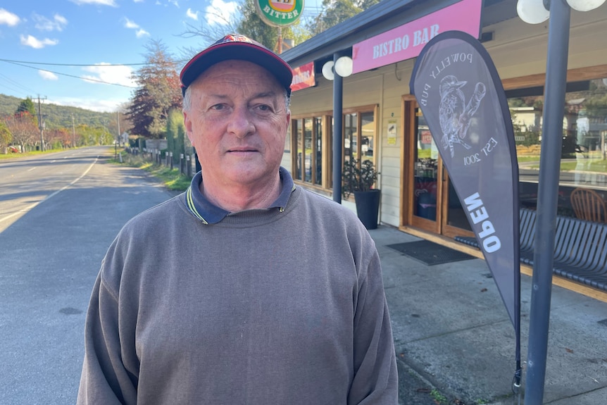 An older man in a cap and a jumper stands outside a pub in a country town.